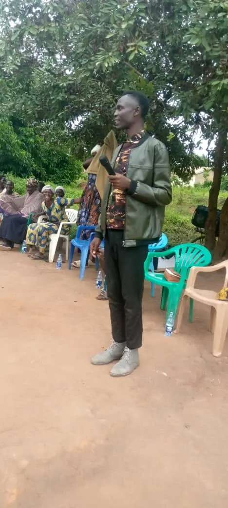 Evangelist holding a microphone preaching to seated village elders under trees